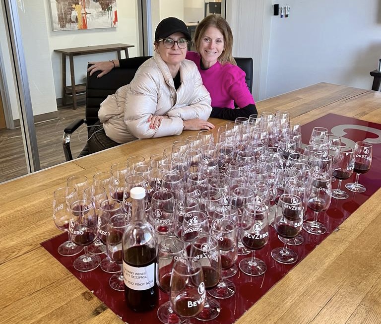 Two women sitting next to each other at a table with dozens of wine glasses resting on it, each with a bit of red wine.