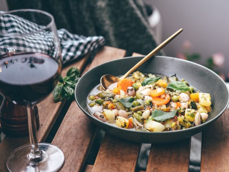 An outdoor table with a bowl of cooked vegetables, a glass of red wine, bread, and salt and pepper shakers on it.