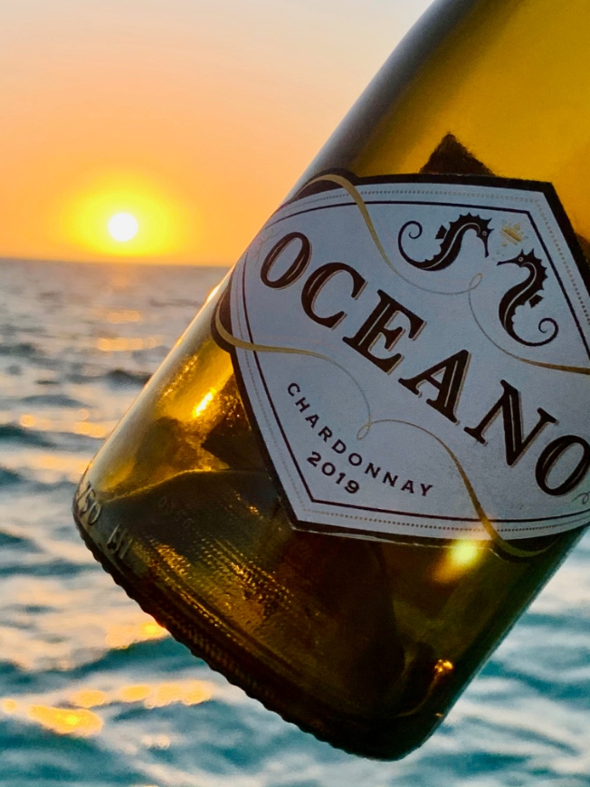 A bottle of oceano chardonnay held up to ocean and sunset.